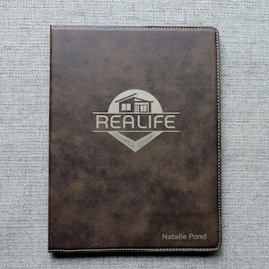May include: A brown leather journal with the words "REALIFE SUSTAINABLE LUXURY" and a house logo. The name "Natalie Pond" is printed at the bottom. The journal has a stitched border.