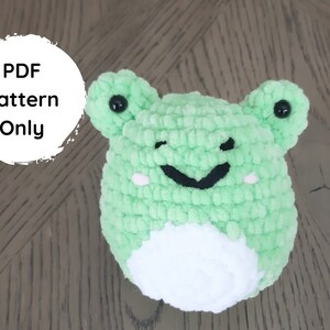 Puede incluir: Un juguete de amigurumi de rana de crochet verde con ojos negros y vientre blanco. El juguete está sentado sobre una superficie de madera. Patrón PDF solamente.