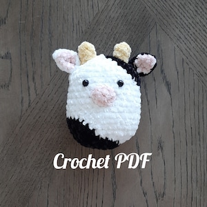 May include: A crocheted stuffed cow toy with black and white fur, yellow ears, and pink nose and hooves. The toy is sitting on a wooden surface. Crochet PDF.