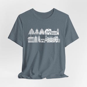 May include: A heathered slate blue short-sleeve t-shirt featuring a white graphic print of two rows of various international architectural landmarks, including towers, pagodas, and historic buildings arranged in a horizontal skyline pattern.
