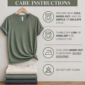 May include: A sage green t-shirt hanging on a wooden hanger with a knot at the hem, stacked with three folded t-shirts in cream, green, and charcoal. The image displays Bella+Canvas 3001 care instructions including machine wash cold, tumble dry low, and cool iron.