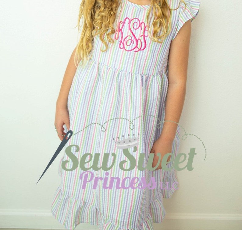 May include: A white dress with multi-colored stripes and a pink embroidered monogram. The dress has a ruffled hem and short sleeves. The dress is made by Sew Sweet Princess.