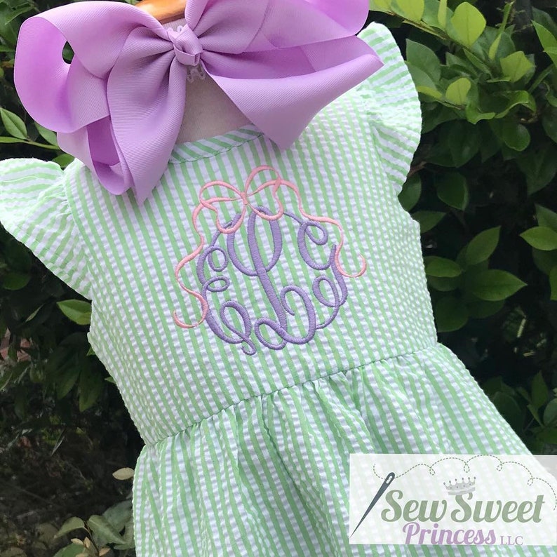 May include: A green and white striped dress with a purple monogram and a pink bow. The dress has ruffled sleeves and a gathered skirt.