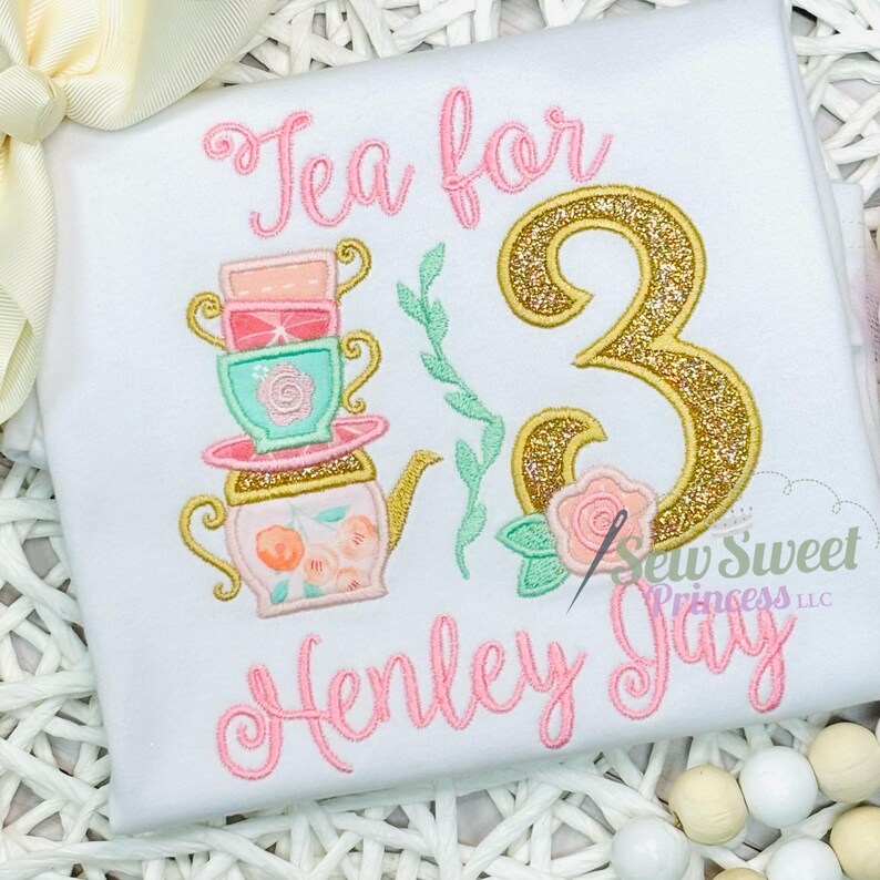 K&ouml;nnte beinhalten: Wei&szlig;es T-Shirt mit einem rosa Aufdruck "Tea for 3 Henley Jay". Das Design umfasst einen Stapel Teetassen, eine Teekanne, eine goldglitzernde Zahl 3 und florale Akzente. Der Text "Sew Sweet Princess LLC" ist ebenfalls sichtbar.