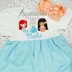 May include: A white and light blue dress featuring embroidered cartoon princesses. The dress has a white top with puff sleeves and a light blue skirt. The text "Sew Sweet Princess LLC" and "Elodie" are also embroidered on the dress. A peach bow is on the top right.