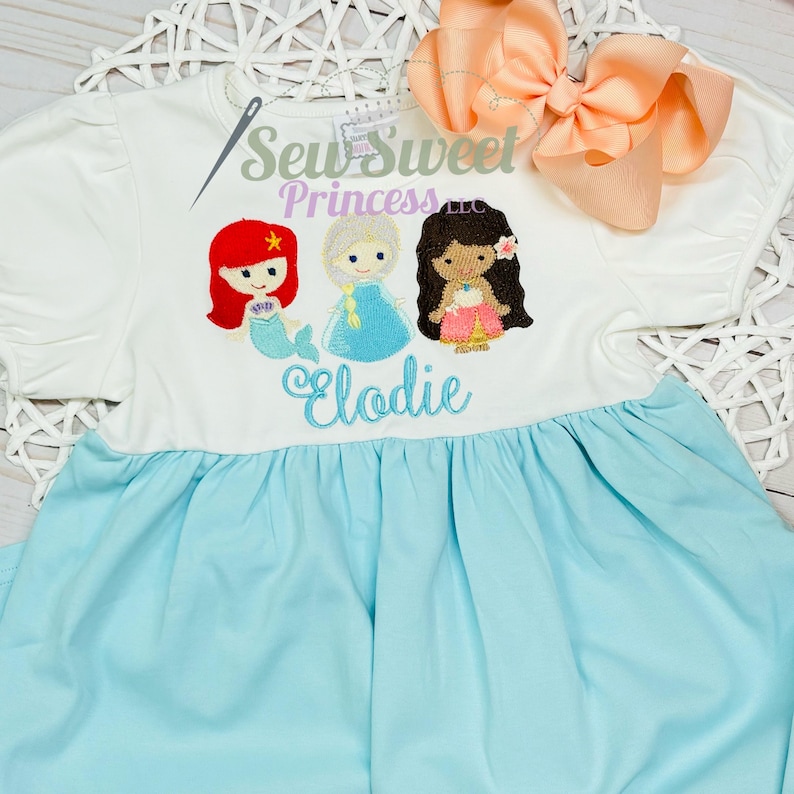 May include: A white and light blue dress featuring embroidered cartoon princesses. The dress has a white top with puff sleeves and a light blue skirt. The text "Sew Sweet Princess LLC" and "Elodie" are also embroidered on the dress. A peach bow is on the top right.