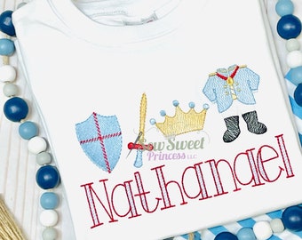 Peut inclure: Combinaison blanche pour bébé avec le texte "Nathaniel" brodé en rouge et des images brodées d'une épée, d'un bouclier, d'une couronne et d'un prince en costume bleu avec des bottes noires.