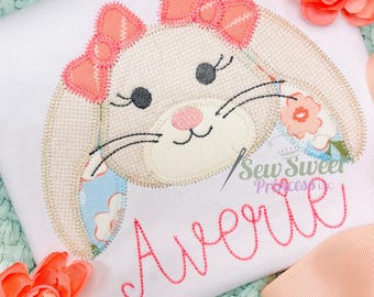 Peut inclure: T-shirt blanc avec un visage de lapin brodé, un nœud rose et des motifs floraux. Le nom "Averie" est brodé en rose. Le design est probablement pour les enfants.