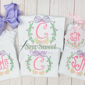 Op de afbeelding: Drie witte shirts met geborduurde bloemkransen en konijnenontwerpen. De kransen hebben een roze strik en een gemonogrammeerde initial in een roze lettertype. De shirts liggen op een witte houten achtergrond.