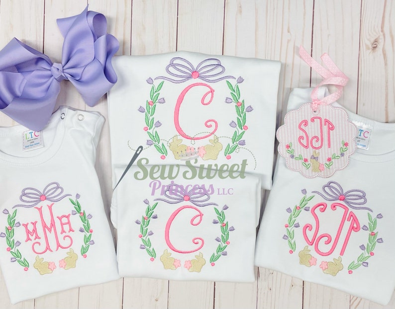 Op de afbeelding: Drie witte shirts met geborduurde bloemkransen en konijnenontwerpen. De kransen hebben een roze strik en een gemonogrammeerde initial in een roze lettertype. De shirts liggen op een witte houten achtergrond.