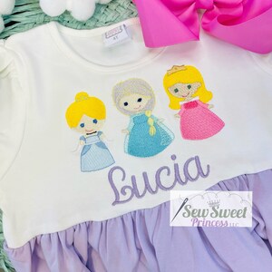 May include: White long-sleeved dress with embroidered cartoon princess figures. The dress features a lilac ruffle hem and the name "Lucia" embroidered in cursive. A pink bow is visible in the background.