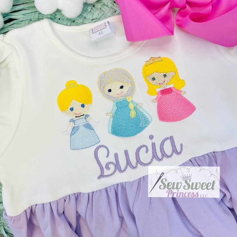 May include: White long-sleeved dress with embroidered cartoon princess figures. The dress features a lilac ruffle hem and the name "Lucia" embroidered in cursive. A pink bow is visible in the background.