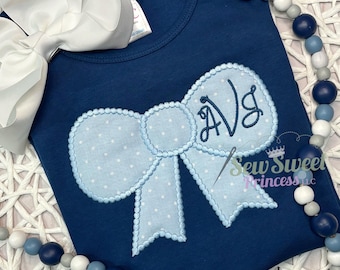 Peut inclure: T-shirt bleu marine avec un nœud papillon bleu clair à pois blancs. Le nœud a un bord festonné et des initiales brodées. Un grand nœud blanc est visible dans le coin supérieur gauche. Le texte "Sew Sweet Princess llc" est visible.