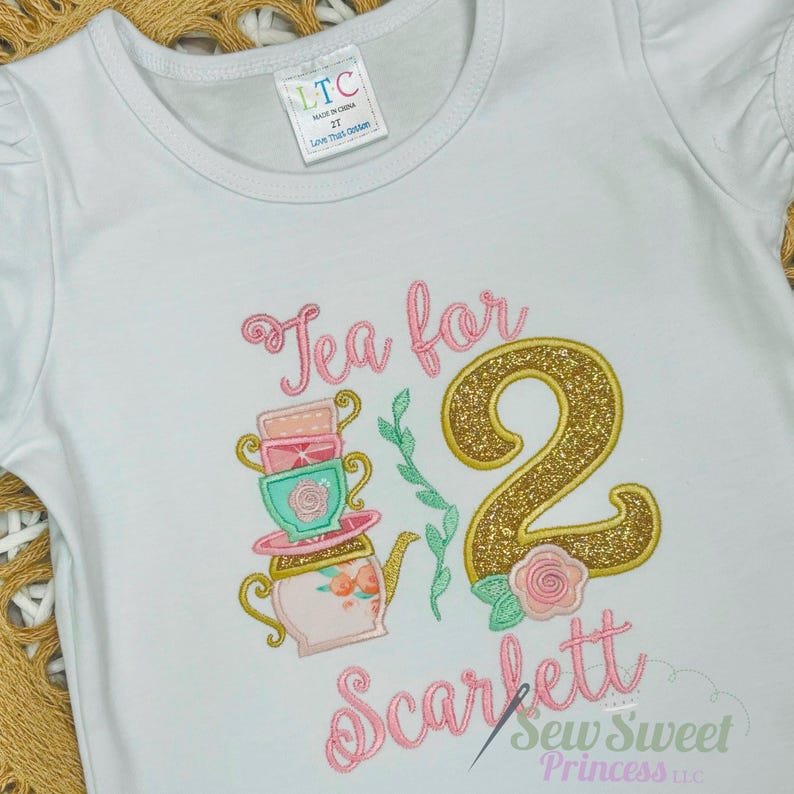 K&ouml;nnte beinhalten: Wei&szlig;es kurz&auml;rmeliges T-Shirt mit der gestickten Aufschrift "Tea for 2 Scarlett" in rosa Schrift. Das Design umfasst einen Stapel Teetassen, eine Teekanne, eine goldene Glitzer "2" und eine Rose. Das Shirt ist mit "LTC Made in China 2T Love This Outfit" gekennzeichnet.