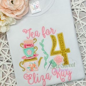 K&ouml;nnte beinhalten: Wei&szlig;es T-Shirt mit einem farbenfrohen Stickdesign. Das Design zeigt eine Teekanne, Teetassen, die Zahl 4 und den Text "Tea for Eliza Kay". Das Design ist in Rosa, Gold und Mintgr&uuml;n gehalten.