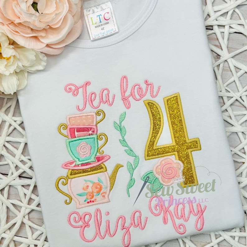 K&ouml;nnte beinhalten: Wei&szlig;es T-Shirt mit einem farbenfrohen Stickdesign. Das Design zeigt eine Teekanne, Teetassen, die Zahl 4 und den Text "Tea for Eliza Kay". Das Design ist in Rosa, Gold und Mintgr&uuml;n gehalten.