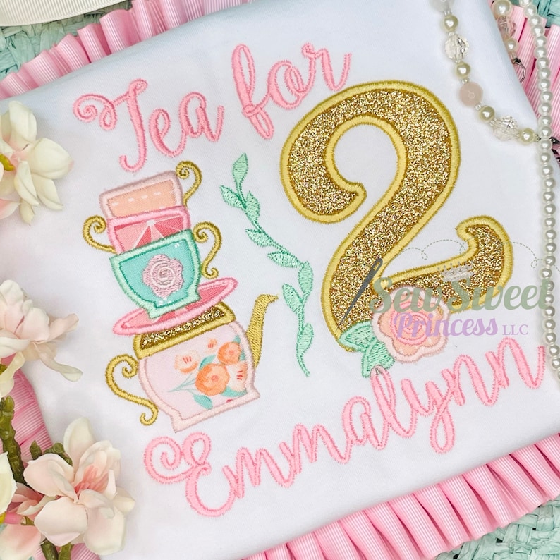 K&ouml;nnte beinhalten: Wei&szlig;es T-Shirt mit rosa gesticktem Text "Tea for 2 Emmalynn". Mit einer goldglitzernden Zahl 2, einer Teekanne und gestapelten Teetassen in Rosa, Gold und T&uuml;rkis. Florale Akzente runden das Design ab.