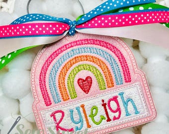Peut inclure: Un porte-clés brodé rose avec un motif arc-en-ciel et le nom "Ryleigh" en fil coloré. Le porte-clés est orné d'un cœur rouge et attaché à un ruban à pois bleus, roses et verts.