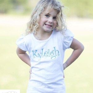 Op de afbeelding: Een wit T-shirt met korte mouwen met een blauwe geborduurde naam "Kyleigh" op de voorkant.