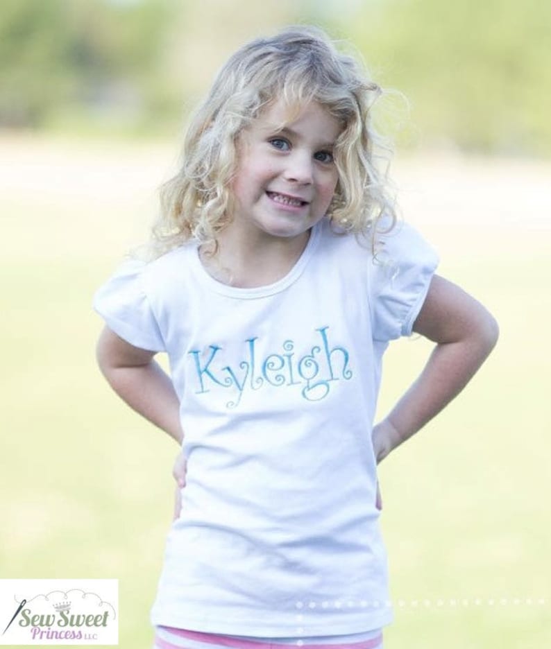 Op de afbeelding: Een wit T-shirt met korte mouwen met een blauwe geborduurde naam "Kyleigh" op de voorkant.
