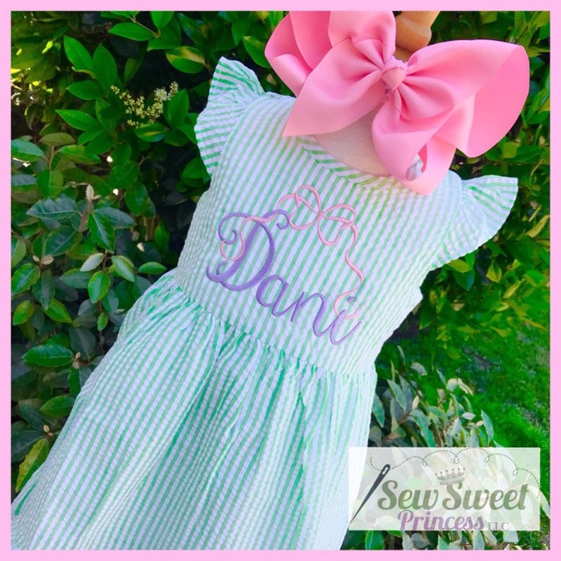 May include: A green and white striped dress with a purple embroidered name "Dani" on the front. The dress has ruffled sleeves and a pink bow on the top.