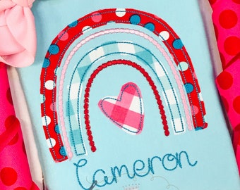 Peut inclure: Un t-shirt bleu clair avec un arc-en-ciel rouge, rose et bleu avec un cœur à carreaux rose et blanc au centre. Le nom "Cameron" est brodé en cursive bleue sous l'arc-en-ciel. Le texte "Sew Sweet Princess LLC" est brodé en bleu sous le nom.