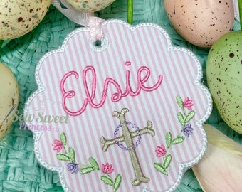Peut inclure: Une étiquette en tissu rayée rose et blanche avec un bord festonné. L'étiquette est brodée du nom "Elsie" en fil rose, d'une croix argentée avec un cercle violet et d'un motif floral.