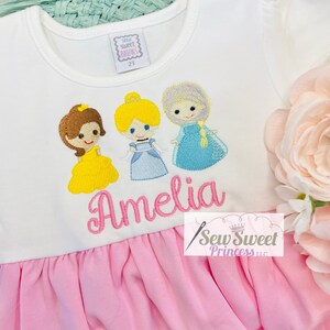 May include: White and pink dress featuring embroidered cartoon princesses. The dress has a white top with the name "Amelia" embroidered in pink, and a pink ruffled skirt. The top also has the text "Sew Sweet Princess llc" and a size tag that says "2T".