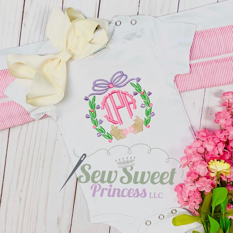 Op de afbeelding: Witte baby romper met een geborduurd monogram in roze, paars en groen en een konijnenontwerp. De romper heeft de tekst "Sew Sweet Princess LLC" onderaan.