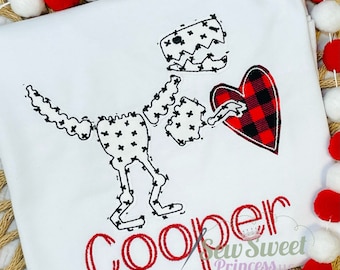 Peut inclure: Un t-shirt blanc avec un squelette de dinosaure noir et blanc tenant un cœur en tissu à carreaux rouge et noir. Le nom "Cooper" est brodé en rouge sous le dinosaure.