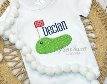Peut inclure: Combinaison blanche pour bébé avec un terrain de golf vert et un drapeau rouge brodés sur le devant. Le nom "Declan" est brodé en bleu au-dessus du terrain de golf.