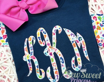 Peut inclure: T-shirt bleu marine avec un grand monogramme coloré. Le monogramme est dans une police fantaisie avec un motif floral rose, jaune, bleu et orange. Un grand nœud rose est attaché. Le texte "Sew Sweet Princess LLC" est en bas.