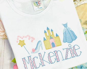 Peut inclure: T-shirt blanc avec un motif brodé rose et bleu représentant un château, une pantoufle de verre, une baguette magique et le nom "Mckenzie".