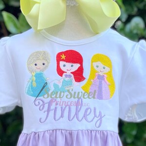 May include: White dress with embroidered images of three cartoon princesses: Elsa, Ariel, and Rapunzel. The dress has a lavender skirt and a large yellow bow. The text "Sew Sweet Princesses LLC" and a name are embroidered on the dress.