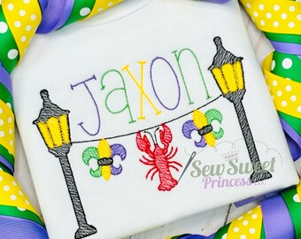 Peut inclure: Body blanc avec le nom "Jaxon" brodé en fil multicolore. Le motif comprend un écrevisse rouge, des fleur-de-lis et des lampadaires. Le body est encadré de rubans colorés.