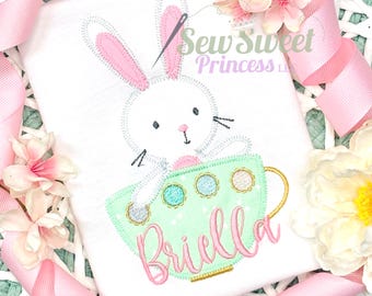 Peut inclure: T-shirt blanc avec un motif brodé représentant un lapin dans une tasse à thé. Le lapin a des oreilles roses et est assis dans une tasse à thé vert menthe à pois colorés. Le nom "Briella" est brodé en rose. Le texte "Sew Sweet Princess LLC" est également visible.