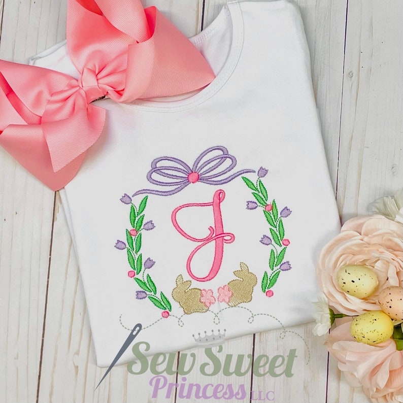 Op de afbeelding: Een witte baby romper met een geborduurde bloemenkrans in roze en paars met een roze letter J in het midden. Twee gouden konijntjes bevinden zich onderaan de krans. De tekst "Sew Sweet Princess LLC" staat onderaan de romper.
