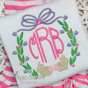 Op de afbeelding: Wit geborduurd shirt met een roze monogram "CRB" omringd door een krans van groene bladeren en paarse bloemen. Twee geborduurde konijnen bevinden zich onder de krans. De tekst "Sew Sweet Princess LLC" staat onderaan het shirt.