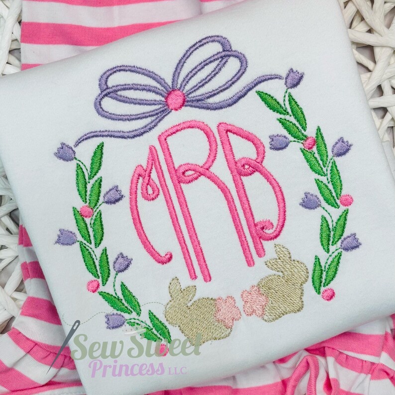 Op de afbeelding: Wit geborduurd shirt met een roze monogram "CRB" omringd door een krans van groene bladeren en paarse bloemen. Twee geborduurde konijnen bevinden zich onder de krans. De tekst "Sew Sweet Princess LLC" staat onderaan het shirt.