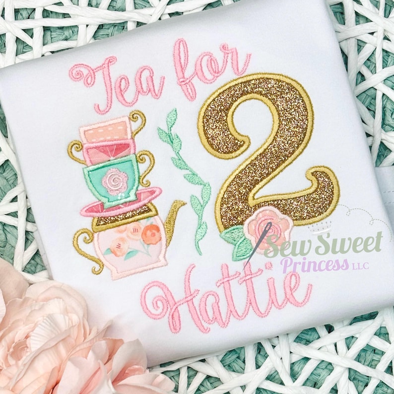 K&ouml;nnte beinhalten: Wei&szlig;es T-Shirt mit gesticktem Design. Mit dem Text "Tea for" &uuml;ber einem Stapel Teetassen und einer Teekanne. Eine gro&szlig;e goldene Glitzer "2" befindet sich neben einer rosa Rose. Der Name "Hattie" ist darunter in rosa Schrift geschrieben.