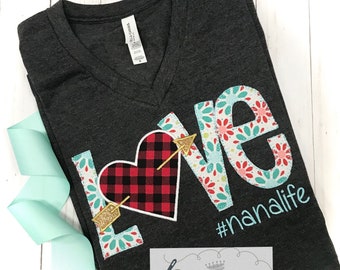 Peut inclure: Un t-shirt gris foncé à col en V avec un motif appliqué coloré qui dit "LOVE" avec un cœur en tissu écossais rouge et noir avec une flèche qui le traverse. Le motif comprend également le texte "#nanalife".