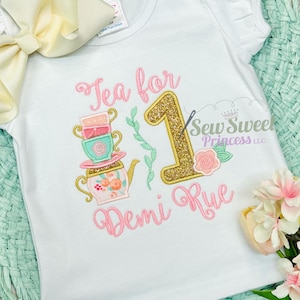 K&ouml;nnte beinhalten: Wei&szlig;es kurz&auml;rmeliges T-Shirt mit einem Teeparty-Design. Das Design enth&auml;lt den Text "Tea for 1 Demi Rue", eine goldglitzernde Zahl 1, einen Stapel Teetassen, eine Teekanne und eine Rose. Das Shirt hat eine hellgelbe Schleife.