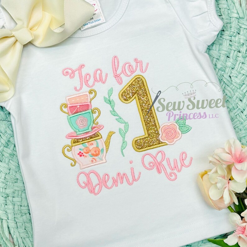 K&ouml;nnte beinhalten: Wei&szlig;es kurz&auml;rmeliges T-Shirt mit einem Teeparty-Design. Das Design enth&auml;lt den Text "Tea for 1 Demi Rue", eine goldglitzernde Zahl 1, einen Stapel Teetassen, eine Teekanne und eine Rose. Das Shirt hat eine hellgelbe Schleife.