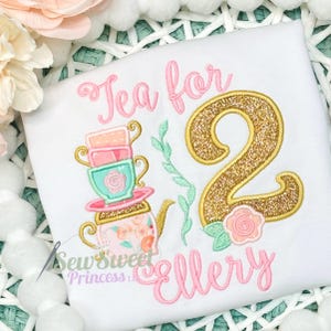 K&ouml;nnte beinhalten: Wei&szlig;es T-Shirt mit rosa gesticktem Text &bdquo;Tea for 2 Ellery&ldquo;. Das Design umfasst einen Stapel Teetassen, eine Teekanne und die Zahl 2 in Goldglitter. Eine rosa Rose und gr&uuml;ne Bl&auml;tter vervollst&auml;ndigen das Design.