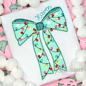 Op de afbeelding: Een wit t-shirt met een geborduurde mintgroene strik met glitterranden en een middenknoop. De strik is versierd met een snoer van kleurrijke kerstverlichting in rood, geel en blauw. De naam Haven is geborduurd in een blauwgroen cursief lettertype.