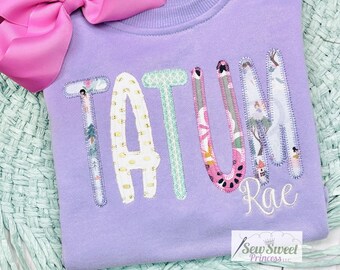 Peut inclure: Sweat-shirt violet avec le nom "TATUM Rae" écrit en lettres de tissu colorées. Les lettres sont décorées de motifs et de designs variés.