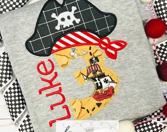 Peut inclure: Un t-shirt gris avec un chapeau de pirate brodé en rouge, un numéro 3 sur le thème de la carte au trésor et le nom "Luke" en broderie rouge.