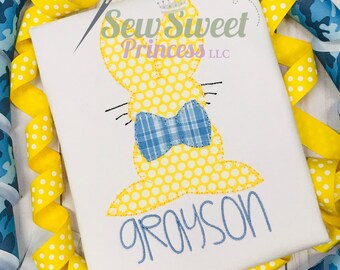 Peut inclure: Un t-shirt blanc avec un lapin jaune portant un nœud papillon bleu et le nom "Grayson" brodé en fil bleu. Le lapin a des pois blancs sur son corps. Le t-shirt est entouré de ruban jaune et bleu.