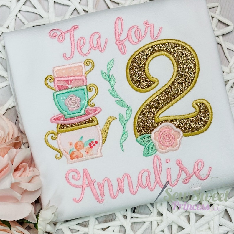 K&ouml;nnte beinhalten: Wei&szlig;es T-Shirt mit einem gestickten Design mit einem Stapel Teetassen, einer Teekanne und dem Text "Tea for 2 Annalise". Die Zahl 2 ist goldglitzernd mit einer rosa Rose und gr&uuml;nen Bl&auml;ttern. Ein s&uuml;&szlig;es Design f&uuml;r einen Geburtstag.