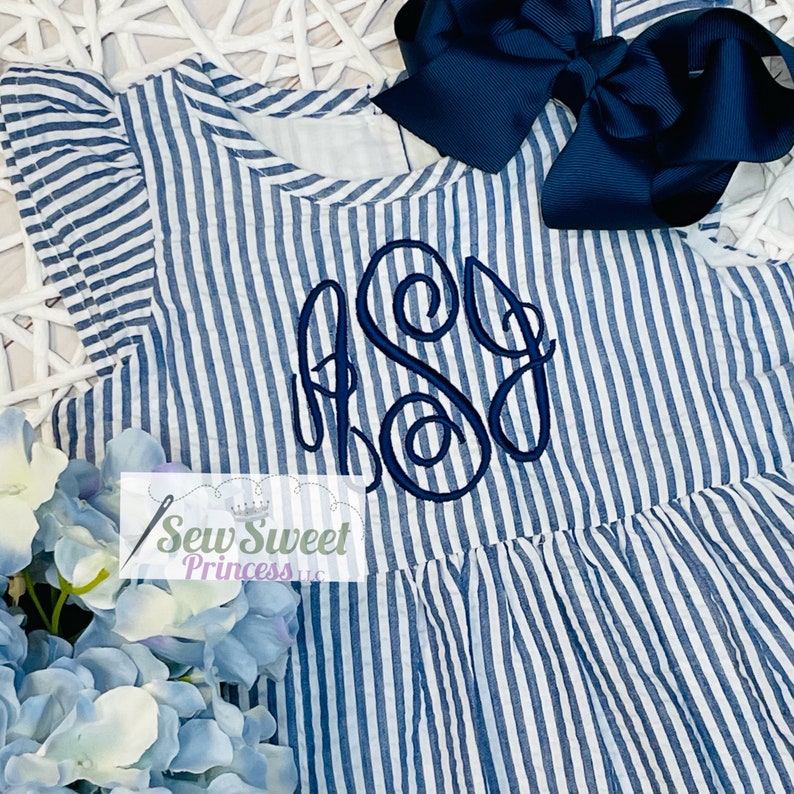 May include: A blue and white striped dress with a ruffled neckline and a monogrammed "SG" on the front. The dress is paired with a navy blue bow.